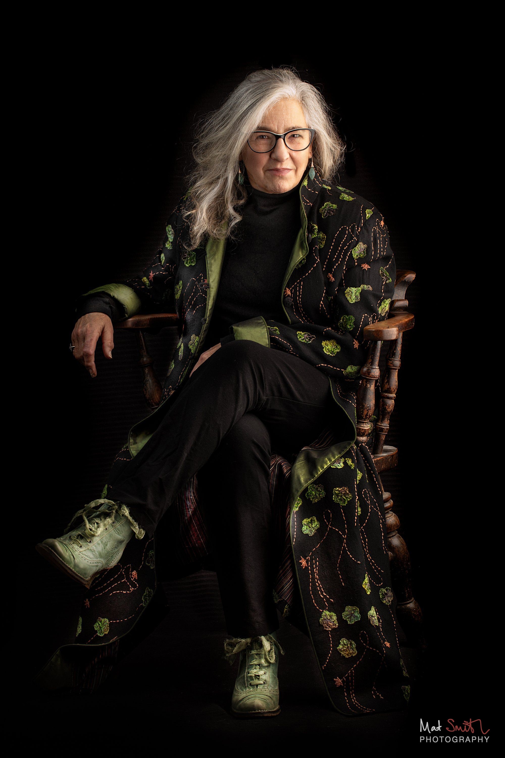 A woman in glasses wearing a long black coat embroidered with green flowers sits in a wooden chair against a black backdrop, portrait by Mat Smith Photography, London