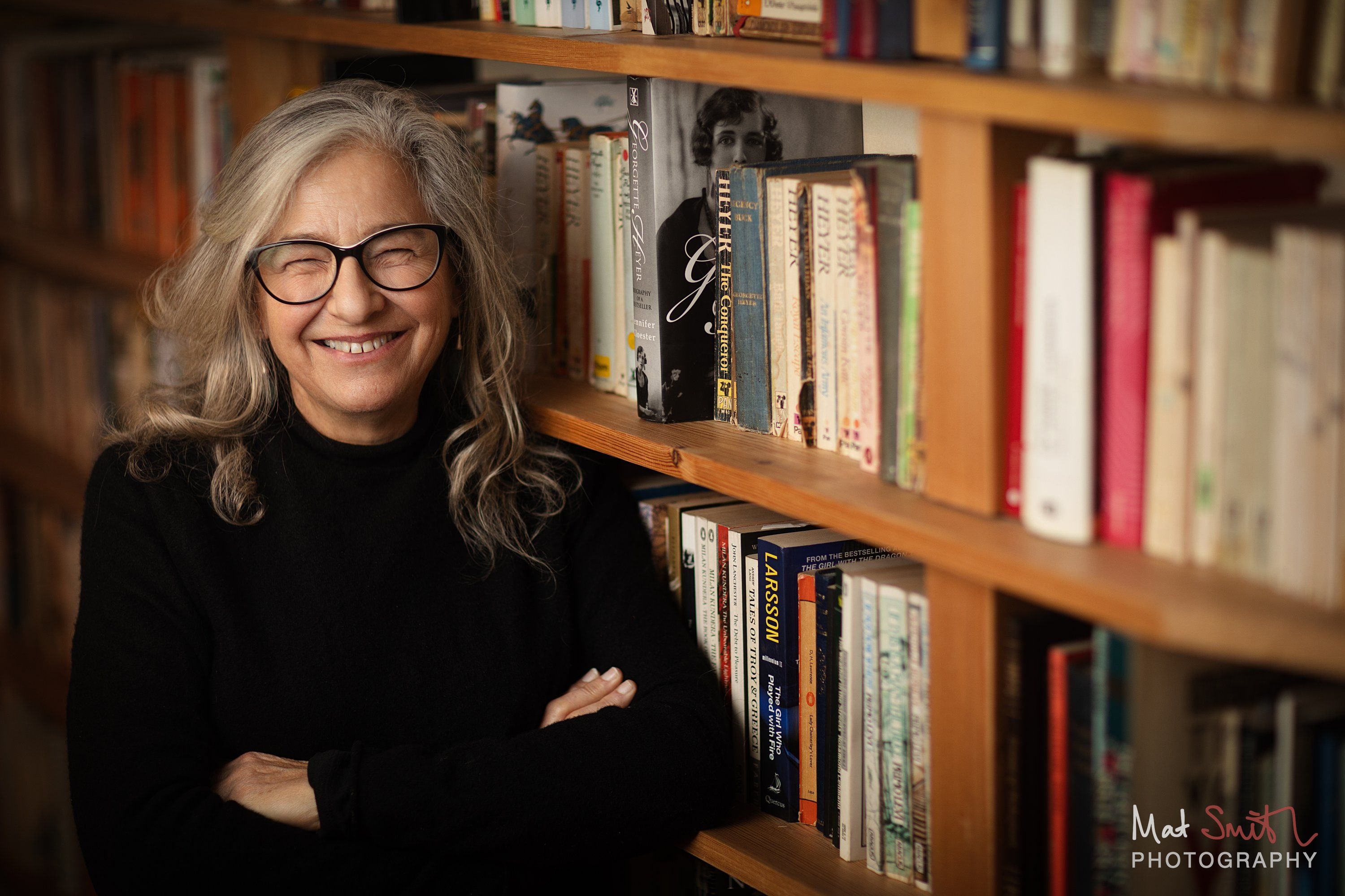A laughing woman with long grey hair and glasses stands with arms folded in front of wooden bookshelves, portrait by Mat Smith Photography, London