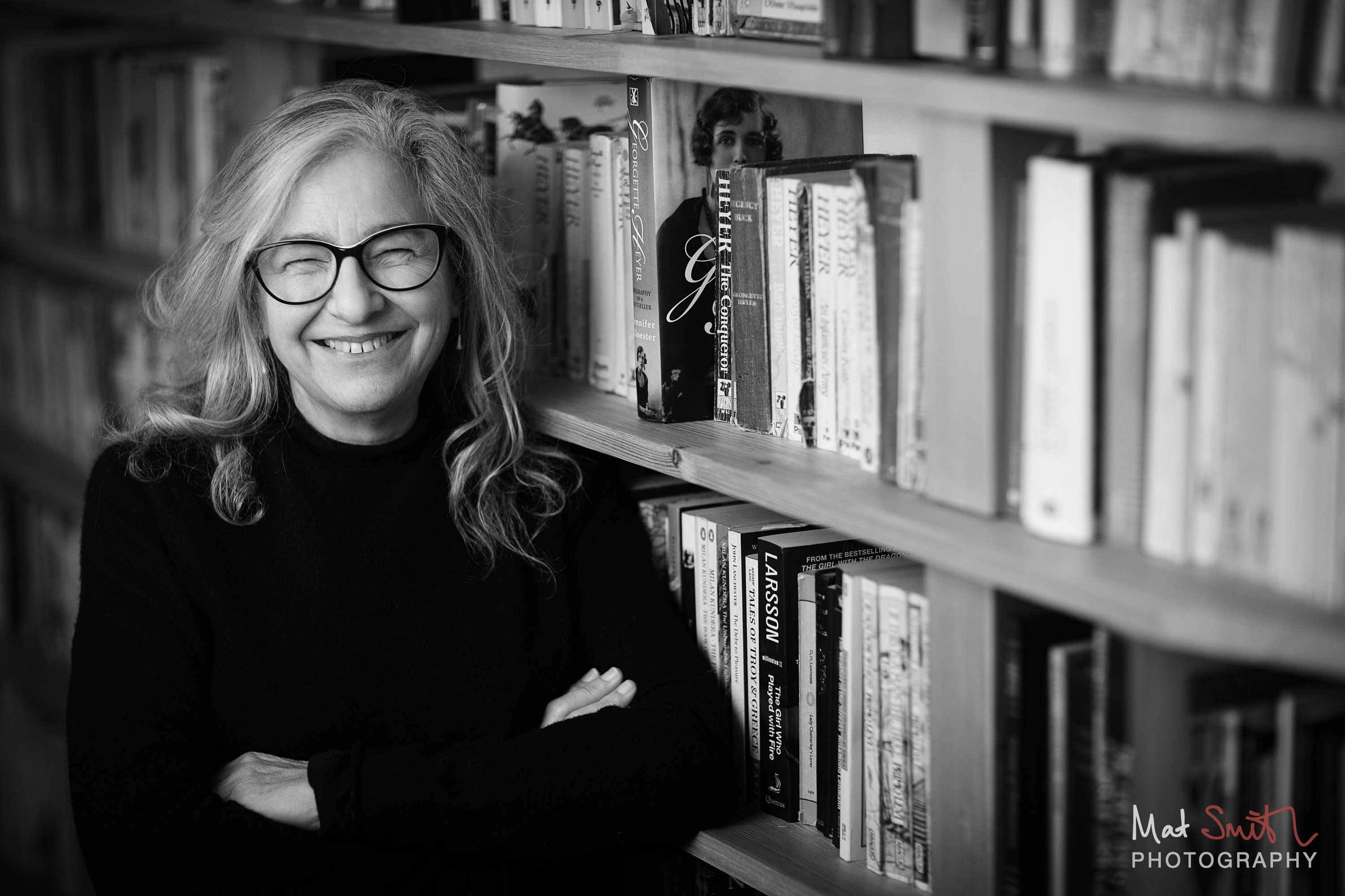 Black and white portrait of a laughing woman in glasses with arms folded in front of bookshelves, portrait by Mat Smith Photography, London