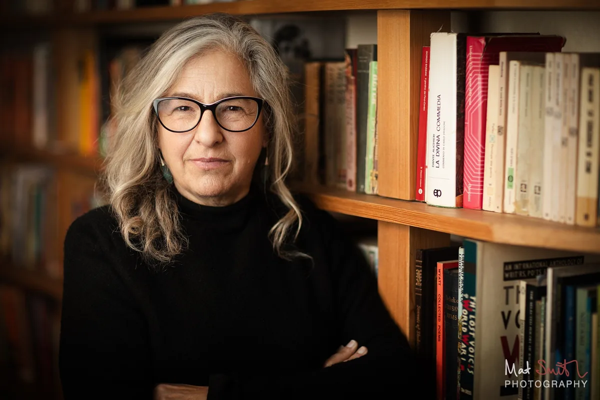 A woman with long grey hair and glasses in a black roll neck looks calmly at the camera in front of full bookshelves, portrait by Mat Smith Photography, London