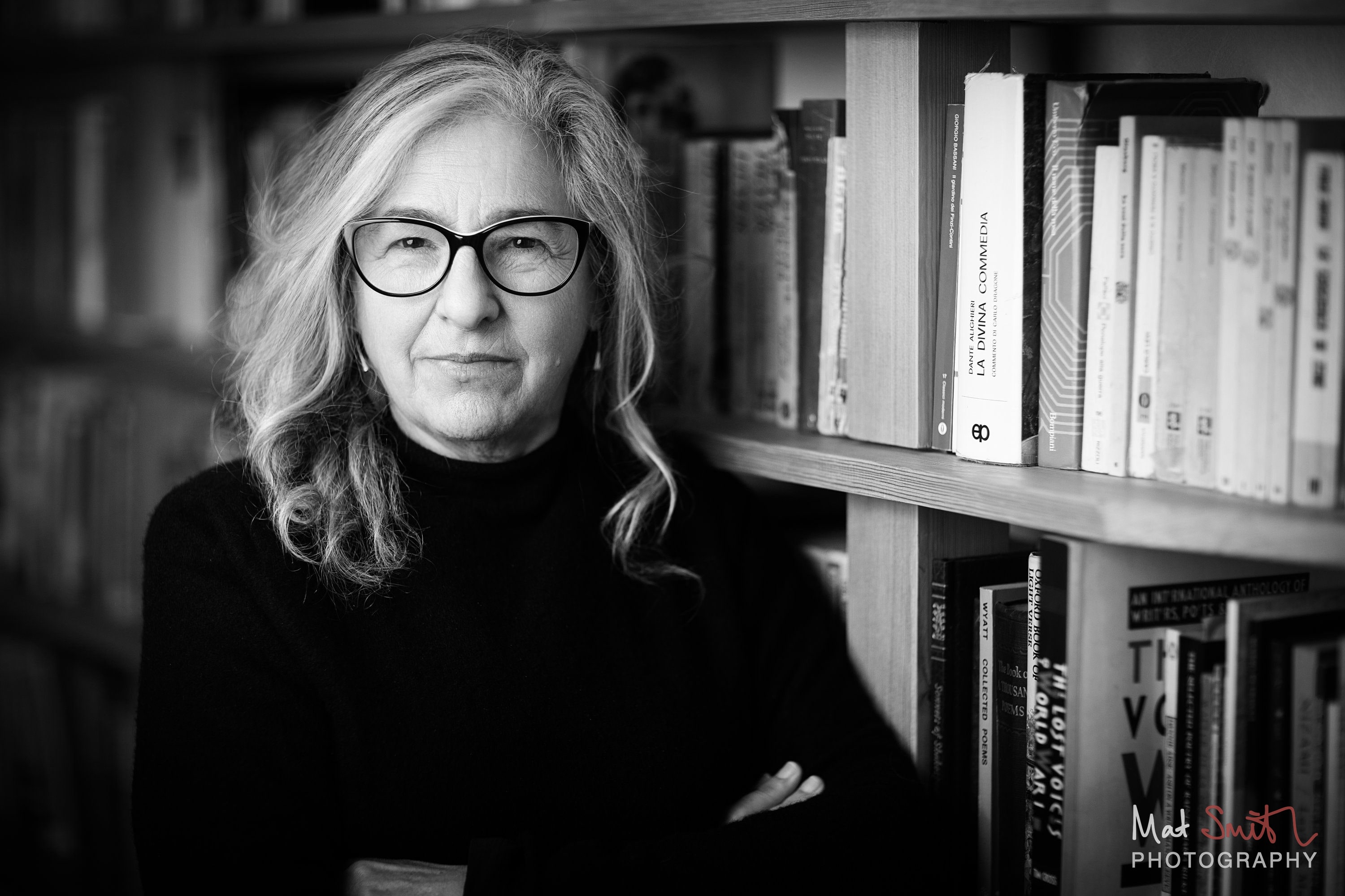 Black and white portrait of a woman in glasses with a calm gaze in front of full bookshelves, portrait by Mat Smith Photography, London