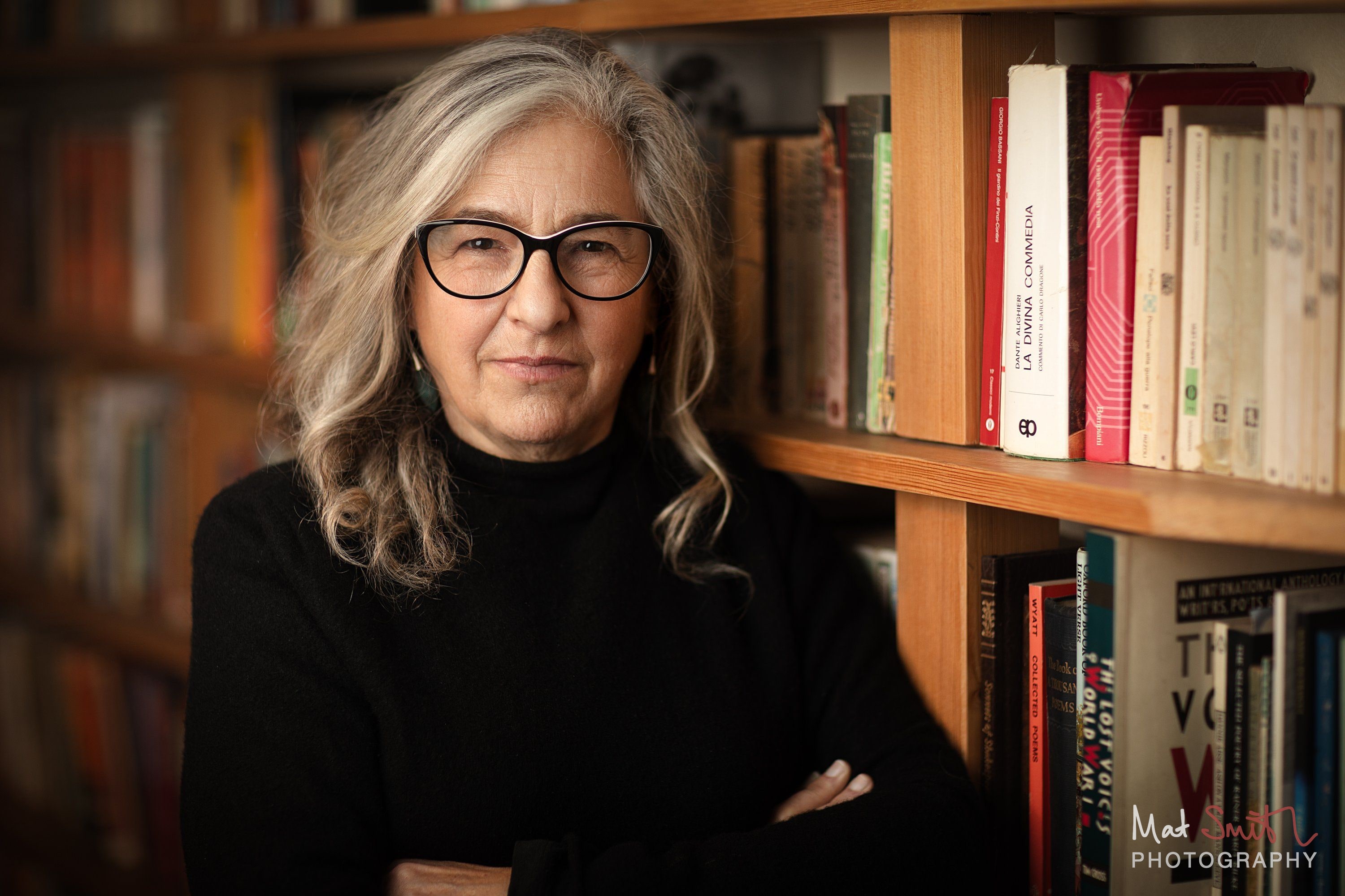 Closer portrait of a woman with long wavy grey hair and glasses with arms folded beside a bookcase in warm light, portrait by Mat Smith Photography, London