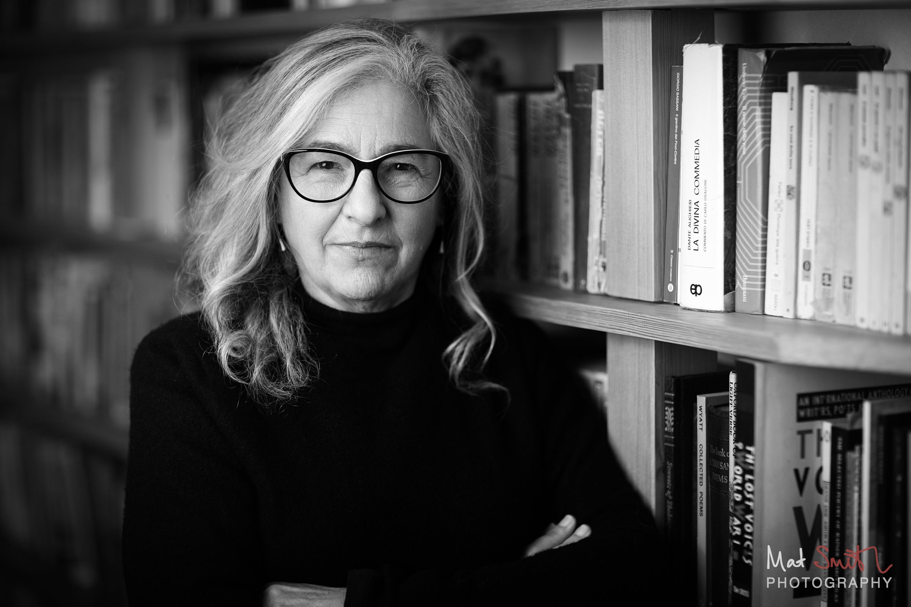 Black and white closer portrait of a woman in glasses with arms folded beside a bookcase, portrait by Mat Smith Photography, London