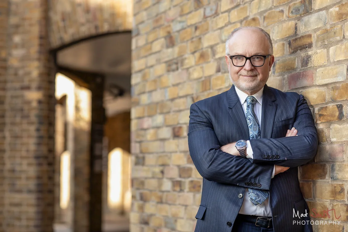 Professor in a navy pinstripe suit and paisley tie, arms folded against a yellow brick wall, portrait by Mat Smith Photography, London