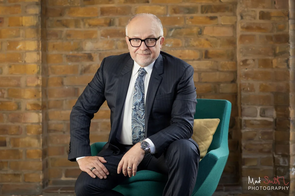 Professor in a navy suit seated on a teal chair in a brick alcove, leaning forward, portrait by Mat Smith Photography, London