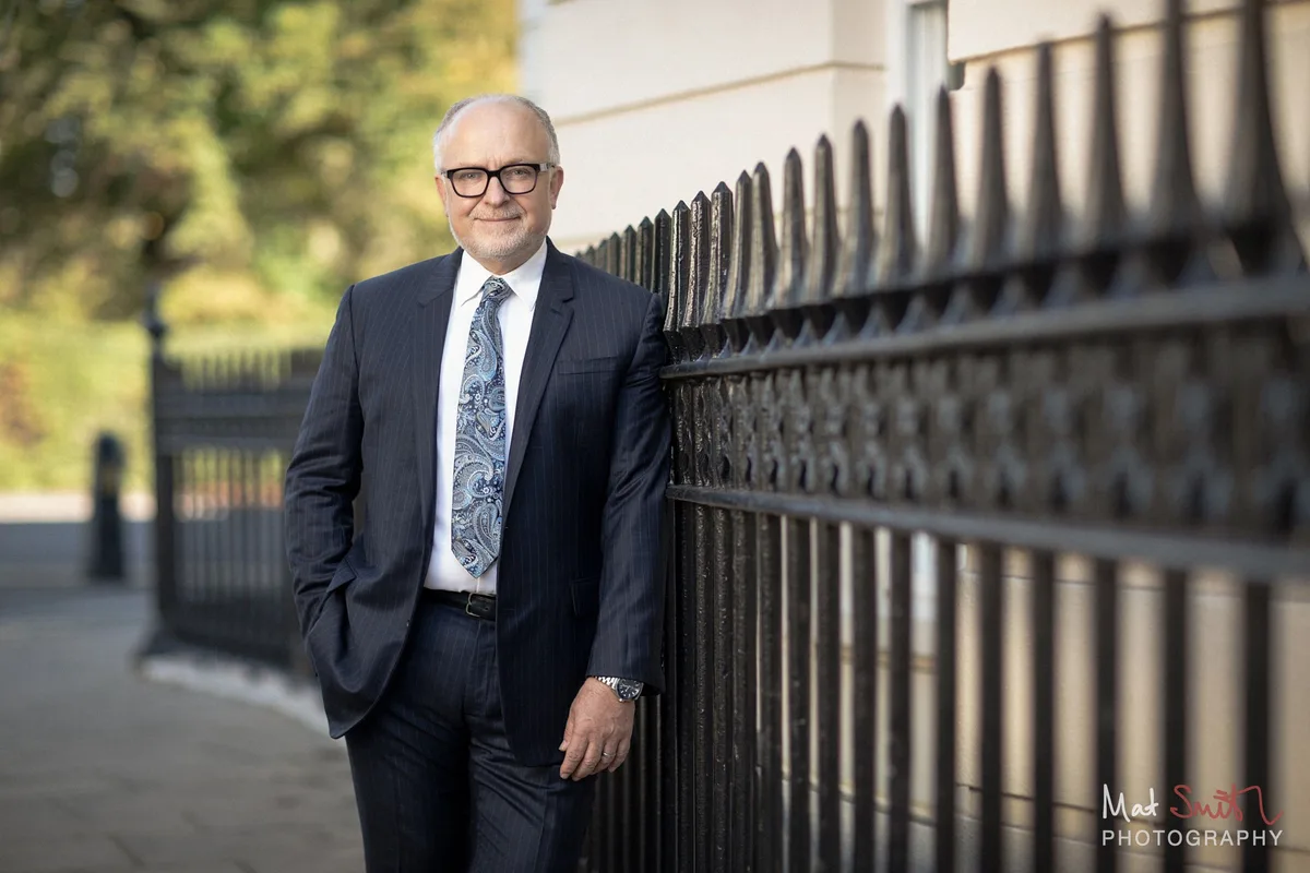 Professor in a navy suit leaning on black iron railings beside a London pavement, portrait by Mat Smith Photography, London