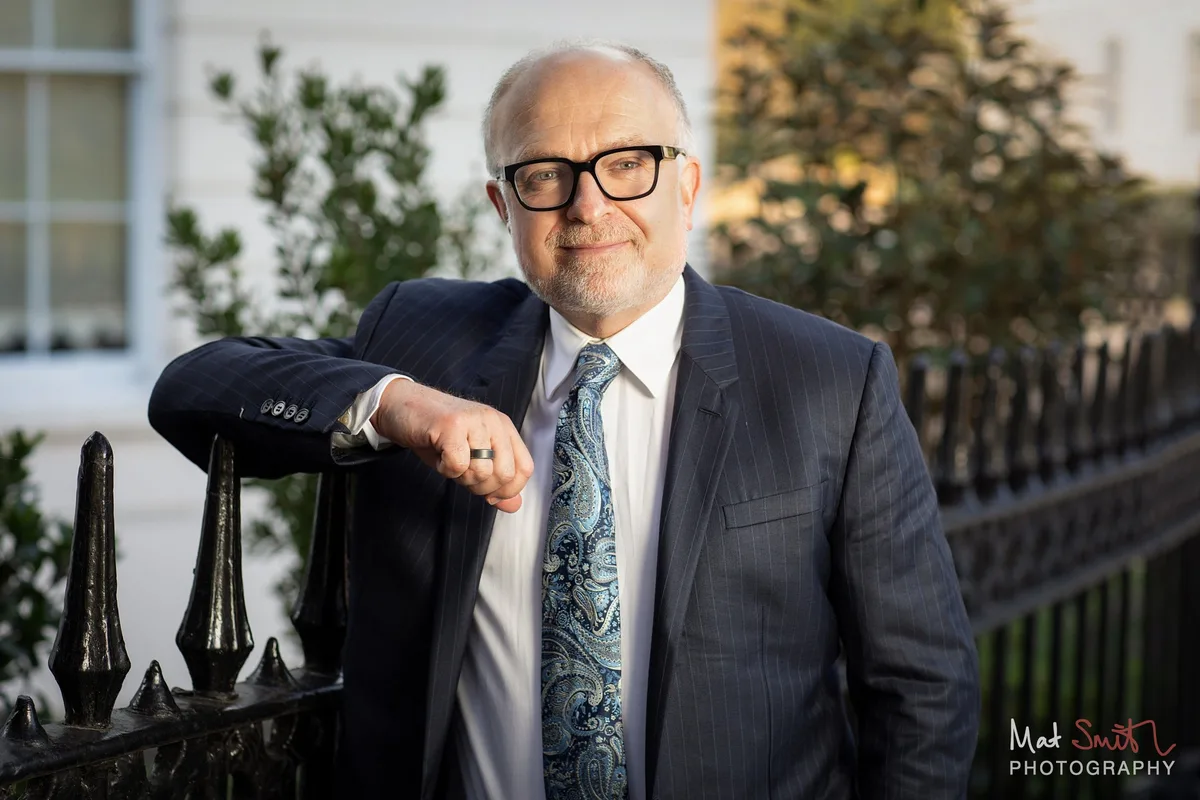 Professor resting an arm on spiked iron railings in front of a white stucco building, portrait by Mat Smith Photography, London