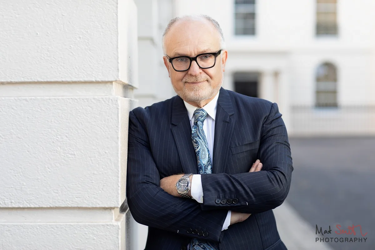 Professor in a navy suit with arms folded beside a white stucco column, portrait by Mat Smith Photography, London