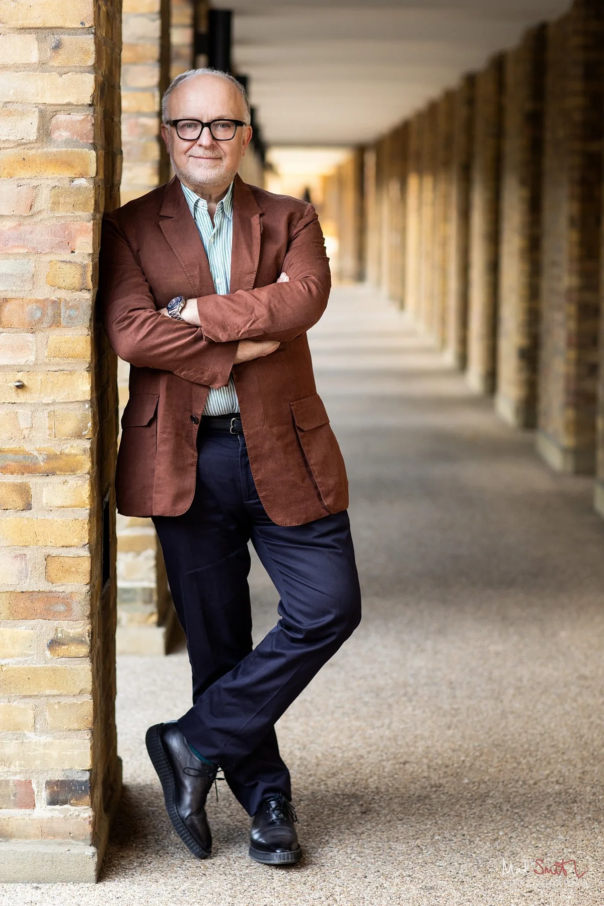 Full length portrait of a professor in a rust jacket leaning in a long brick colonnade, portrait by Mat Smith Photography, London