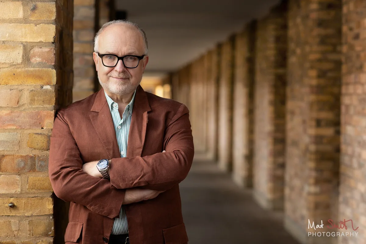 Professor in a rust jacket and striped shirt, arms folded in a brick colonnade, portrait by Mat Smith Photography, London