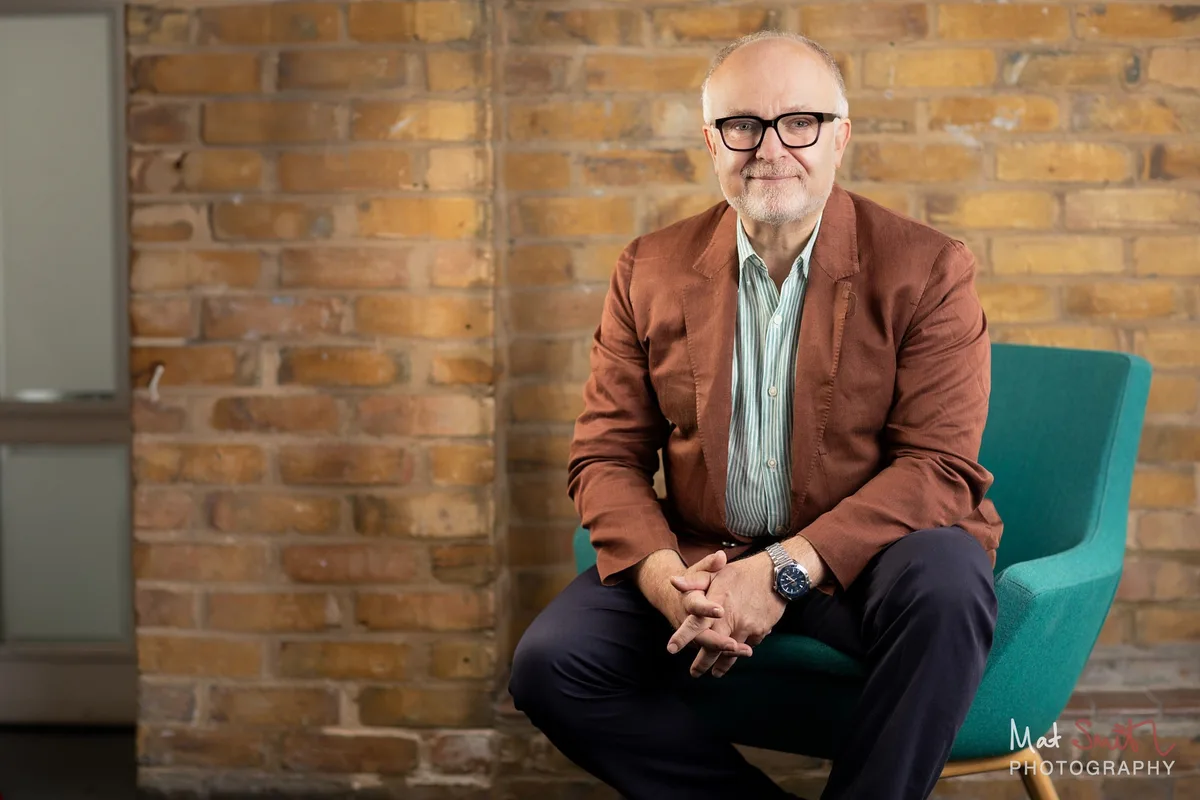 Professor in a rust jacket seated on a teal chair against a brick wall, hands clasped, portrait by Mat Smith Photography, London